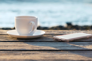 White Cup on black coffee  and mobile on beach at sunrise or sunset for lifestyle and blogger concept. Remote work. Copy space.