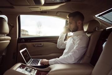 Fototapeta premium Man uses laptop and talking on smartphone sitting in back seat of car, businessman in taxi