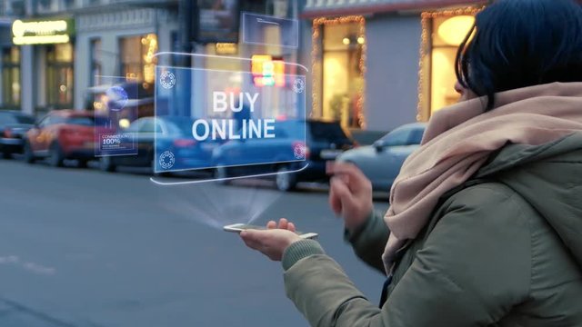 Unrecognizable woman standing on the street interacts HUD hologram with text Buy Online. Girl in warm clothes with a scarf uses technology of the future mobile screen on background of night city