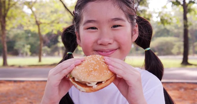 Asian little girl eating hamburger with happy emotion at park. Girl enjoy for eating hamburger alone.
