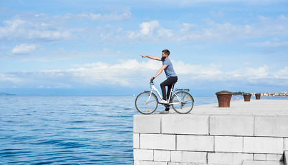Young bearded male biker on bicycle on high paved stone sidewalk pointing at distant mountain on...