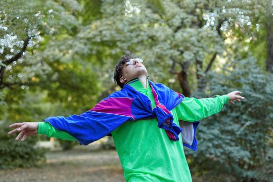 Young handsome caucasian man in bright freaky sportswear in 70s style stands at a morning park, hands widely outstretched. Stick in the mouth, golden glasses, extravagant fashion. Outdoors, copy space