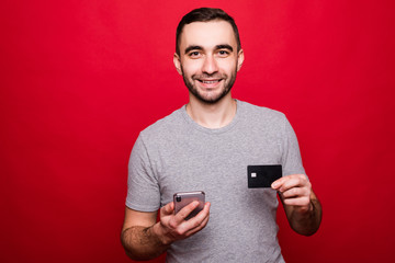 Photo of happy young man isolated over red wall background holding mobile phone and credit card.