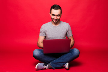 Portrait of a smiling young man using laptop computer while sitting on a floor isolated over red background