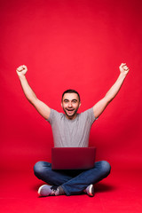 Portrait of a joyful young man looking at laptop computer while sitting on a floor and celebrating isolated over red background