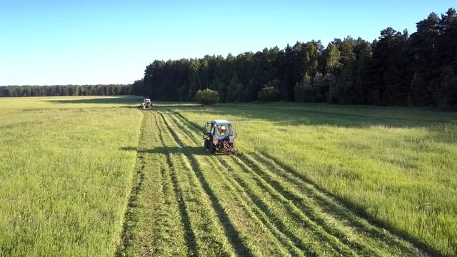 close flight above tractor with mower cutting off thick green hay grass making rolls against forest on hot summer day