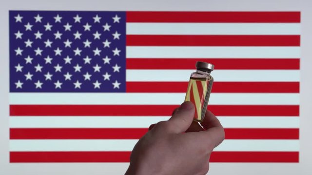 A man holds a glass vial of medicine, United States flag visible on the background.