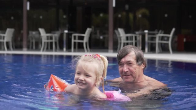 Grandpa is swimming in the pool with his little granddaughter. An elderly man helps a child to float in an outdoor pool. Grandfather and little granddaughter on holiday in Cyprus