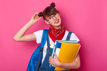 Studio shot of smiling beautiful schoolgirl wearing denim overalls, white t shirt, bandana, touches her bunch. Dark haired student being glad to came back to school
