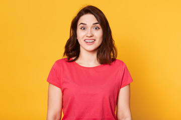Obraz premium Close up portrait of charming brunette lady against yellow studio wall, standing with open mouth, smiling, surprised by pleasant news, wearing red t shirt, copy space for promotion or advertisment.