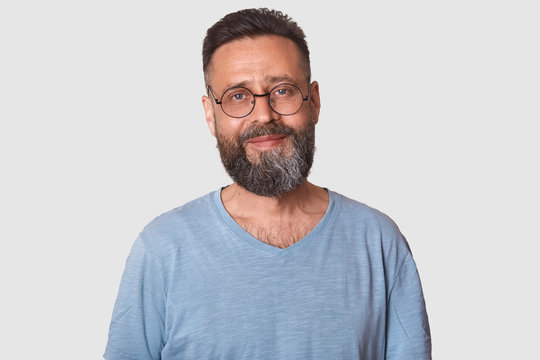 Smiling Handsome Middle Aged Man Posing Against White Background, Looks Cheerful. Black Haired Attractive Model With Beard Wearing Grey T Shirt And Glasses, Has Calm And Pleasant Facial Expression.