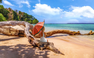 Young female tourist with umbrella sitting on a fallen tree trunk at the scenic Jolly Buoy island sea beach at Andaman islands India
