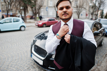 Stylish indian businessman in formal wear tied his tie and standing against black business car on street of city.