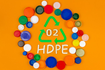 Plastic colorful reusable hdpe bottle caps stacked in circle around recycling sign with code 02. Nature pollution contamination, ecology environmental problem damage concept. Orange background.