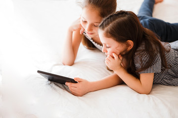 Children using digital gadgets at home. Little girls playing games on the tablet lying on the bed in the bedroom.