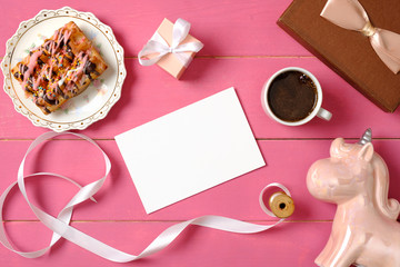 Hipster girls accessories on pink wooden desk: blank paper card, ribbon, unicorn, cup of coffee, gift box, tasty wafer cake. Glamour women desk concept. Flat lay design, top view.