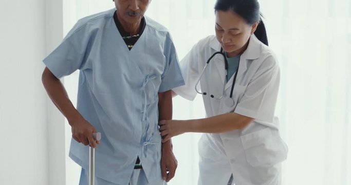 Physiotherapy female doctor helping to support Asian male patient to walking with a cane in hospital.