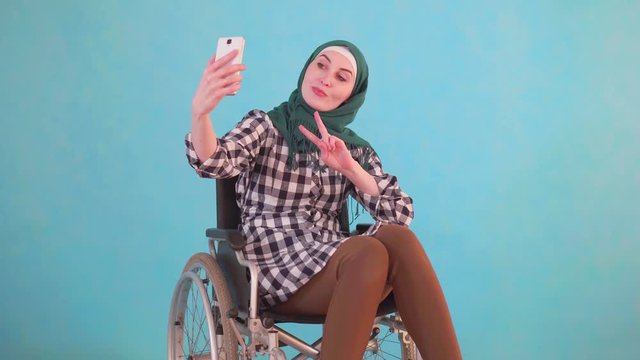 young muslim woman disabled in a wheelchair takes a selfie with her phone on a blue background