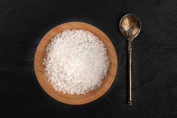 A photo of a bowl of sea salt and a spoon, shot from the top on a black slate background with a place for text