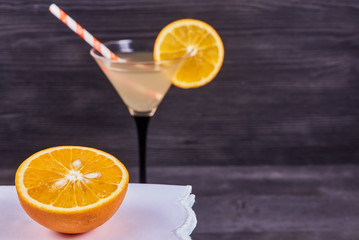 Sliced orange next to martini goblet with fresh orange juice and orange slice, cocktail tube, on a dark wooden background. Focus on orange.
