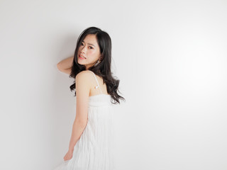Portrait of beautiful Chinese woman in sexy white dress posing with white wall background, young girl looking back and smiling at camera.