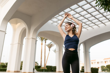 Enjoying lovely music through headphones of positive joyful sportswoman stretching hands above on east architecture background. Fashionable model, having fun. Place for text