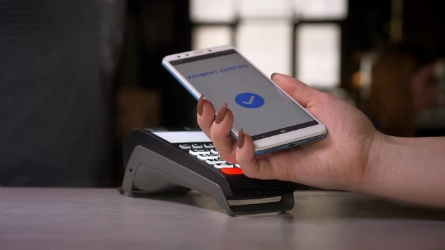Close-up shot of woman attaches smartphone to terminal performing successful contactless payment getting a cup of coffee.