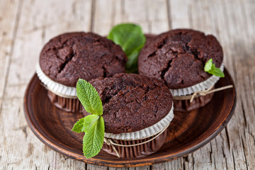 Three chocolate dark muffins with mint leaves on brown ceramic plate.