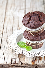 Two fresh dark chocolate muffins with mint leaves on white plate on rustic wooden table.