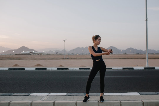 Active joyful sportswoman walking on road on sunset background. Tropical country, looking at watch on hand, cheerful mood, motivation, workout, healthy lifestyle. Place for text