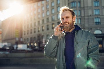 Young bearded man talk on phone outside. Modern buildings behind. Sun shining. Urban view.