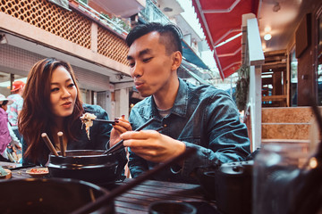 Smiling young chinese couple sitting at restourant outside and trying traditional asian food at...