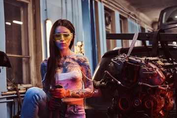 Obraz na płótnie Canvas Female model with tattooed body wearing protective goggles posing with a steel hammer next to a car engine suspended on a hydraulic hoist in the workshop. Photo with red light illumination