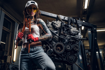 Fototapeta na wymiar Sexual tattoed girl wearing cap and dirty clothes posing next to a car engine suspended on a hydraulic hoist in the workshop