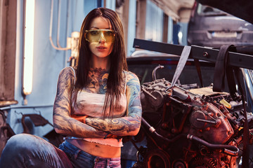 Female car mechanic holding a big hammer and posing next to a car engine suspended on a hydraulic...