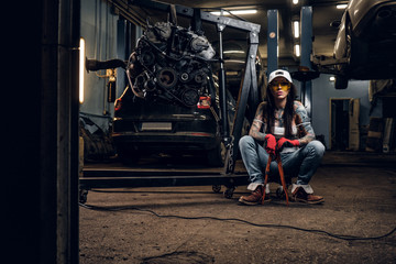 Fototapeta na wymiar Stylish tattooed girl holding a big wrench while sitting on a hydraulic hoist with a suspended car engine in the workshop