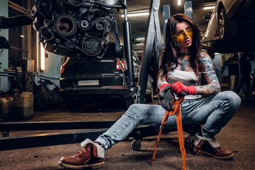 Naklejka na ściany i meble Stylish tattooed girl holding a big wrench while sitting on a hydraulic hoist with a suspended car engine in the workshop