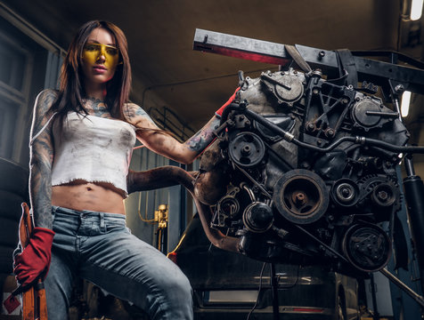 Stylish Tattooed Girl Holding A Big Wrench And Posing Next To A Car Engine Suspended On A Hydraulic Hoist In The Workshop.