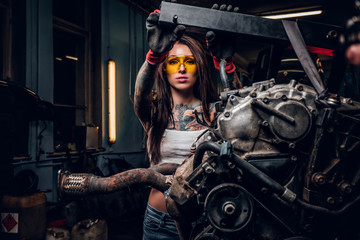 Stylish female model with tattoed body repairs a car engine suspended on a hydraulic hoist in the...