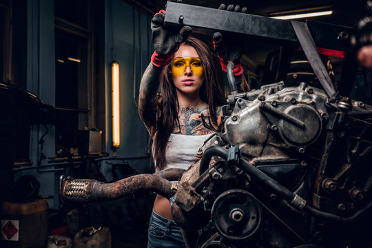 Stylish female model with tattoed body repairs a car engine suspended on a hydraulic hoist in the workshop.
