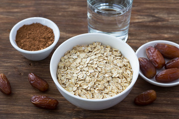 Raw oatmeal in a white bowl, dates fruits, cocoa and a glass of water, ingredients for delicious healthy breakfast