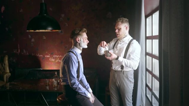 Tattooed barber applies foam by shaving brush on face of his tattooed client, barber shop in 30's mafia style, shaving salon, 4k UHD 60p Prores HQ 422