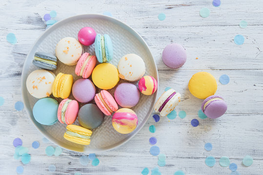 Multicolored french macaron cakes on blue plate and white wooden background. White, yellow, pink, purple and grey french macarons with fresh berries.