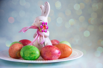 Happy easter bunny. Beautiful easter composition from an Easter rabbit figurine with a pink bow, shiny colored eggs on a blue background. Copy space, bokeh effect