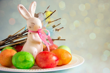 Happy easter bunny. Beautiful easter composition from an Easter rabbit figurine with a pink bow, shiny colored eggs and pussy-willow twigs on a blue background. Copy space, bokeh effect
