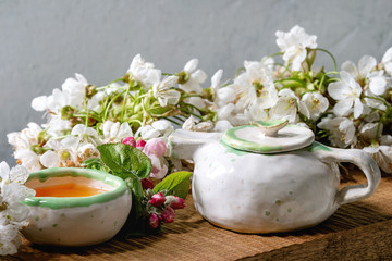 Craft handmade ceramic teapot and cup with hot tea decorated by spring blossom cherry branches on japanese style wooden serving tray on white marble table.