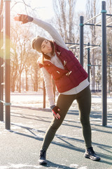 Sports girl in warm clothes on a sunny day does a warm-up on the outdoor sports field in the winter...