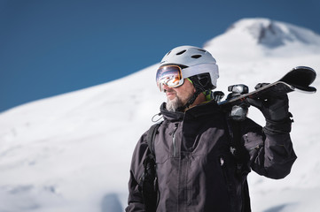 Portrait bearded male skier aged against background of snow-capped Caucasus mountains. An adult man wearing ski googles mask and helmet skis on his shoulder looks mountains. Ski resort concept