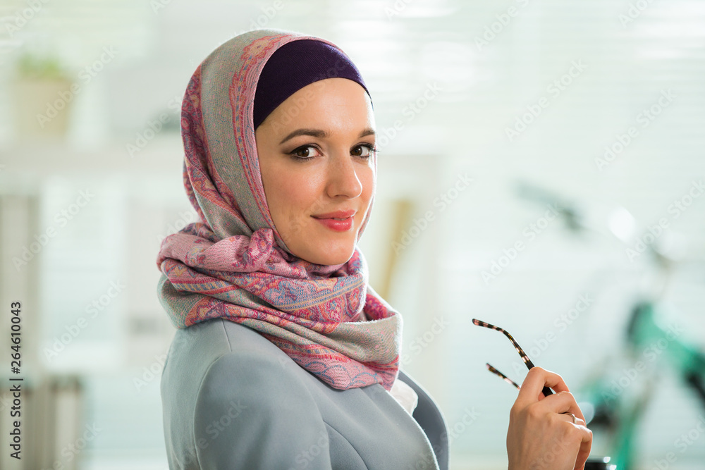 Wall mural Beautiful stylish woman in hijab and eyeglasses, sitting at desk with laptop in office. Portrait of confident muslim businesswoman. Modern office with big window, bicycle on background. 