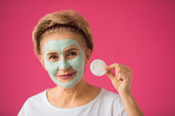 beautiful elderly woman with a wellness cream om face mask on a pink background with a cleansing...
